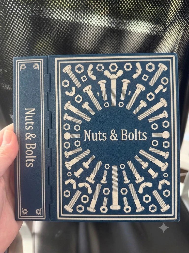 A navy-blue book-shaped box with an embossed silver antique cover reading 'Nuts & Bolts' — a radial sunburst of bolts, nuts and washers around the central title — shown held in one hand against a dark mesh background.