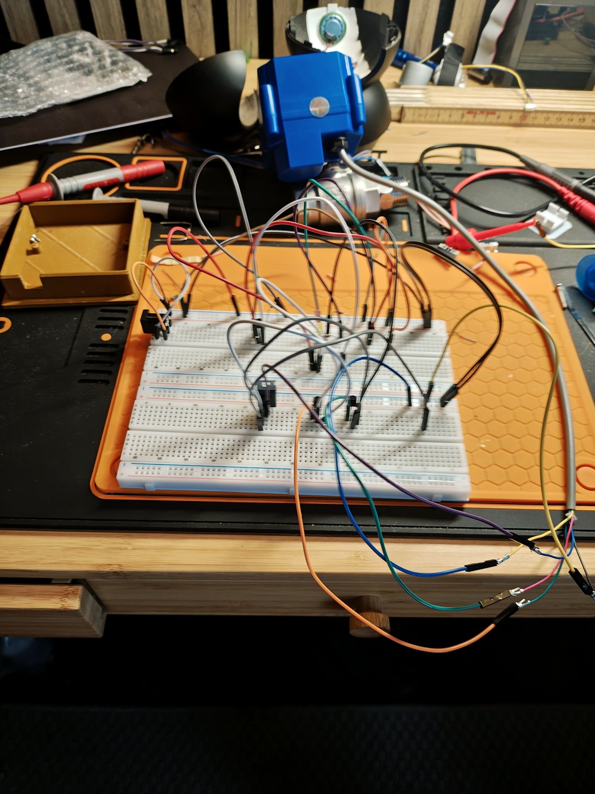 A solderless breadboard wired with a tangle of jumper wires and several small transistors, connected to a blue CWX-15Q motorised ball valve, with multimeter probe leads nearby, sitting on a workbench mat.