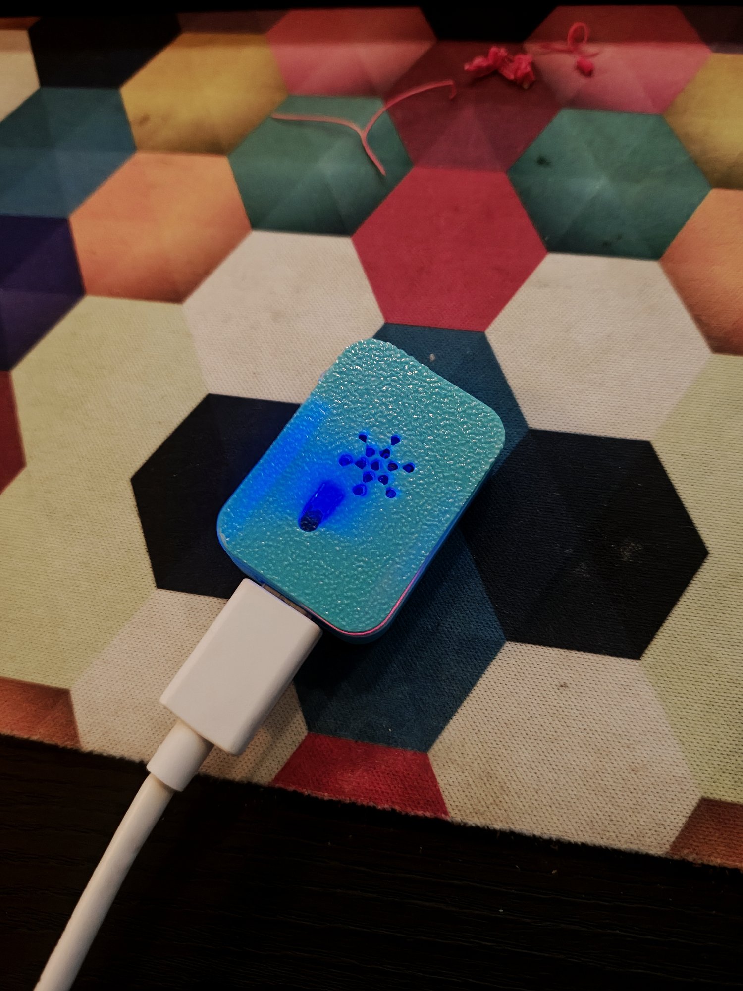 The finished 3D-printed enclosure closed with its lid on, plugged into a white USB-C cable, sitting on the same hexagon-patterned surface. Blue LED light from inside is escaping through the small asterisk-shaped vent pattern in the lid and around the USB-C plug.