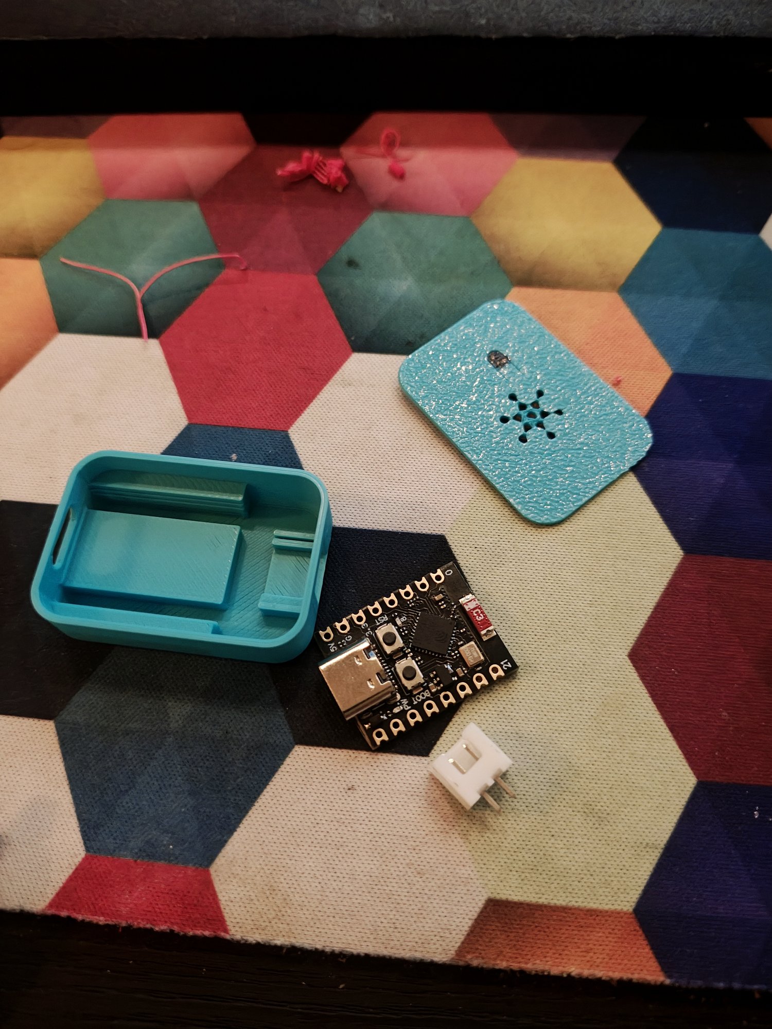 Three things on a colourful hexagon-patterned surface: the two halves of a light-blue 3D-printed enclosure (a lid with a small asterisk-shaped vent pattern, and a tub with mounting pillars inside), and a small ESP32-C3 development board with a USB-C connector and a spare 2-pin connector next to it.
