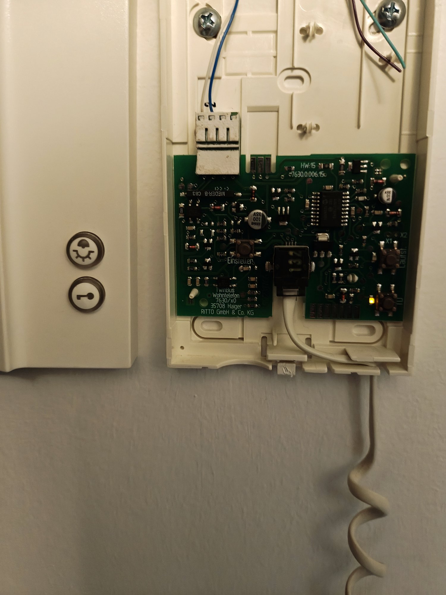 The Ritto TwinBus intercom handset opened on the wall. The top half of a green PCB is visible, with a couple of small tactile buttons near the top edge and a yellow LED lit. Below the PCB is a white bezel with two round buttons: a light-bulb icon and a key icon for door-open.