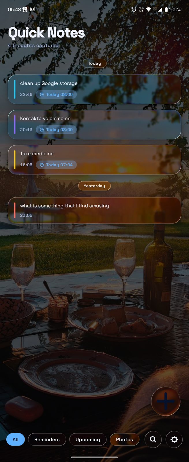 The Quick Notes main screen with a custom photo wallpaper behind it: a sunset outdoor dining table with wine glasses, plates and a wine bottle. Note cards float above the photo with subtle translucent panels. The bottom bar shows Reminders / Upcoming / Photos tabs alongside search and settings icons.