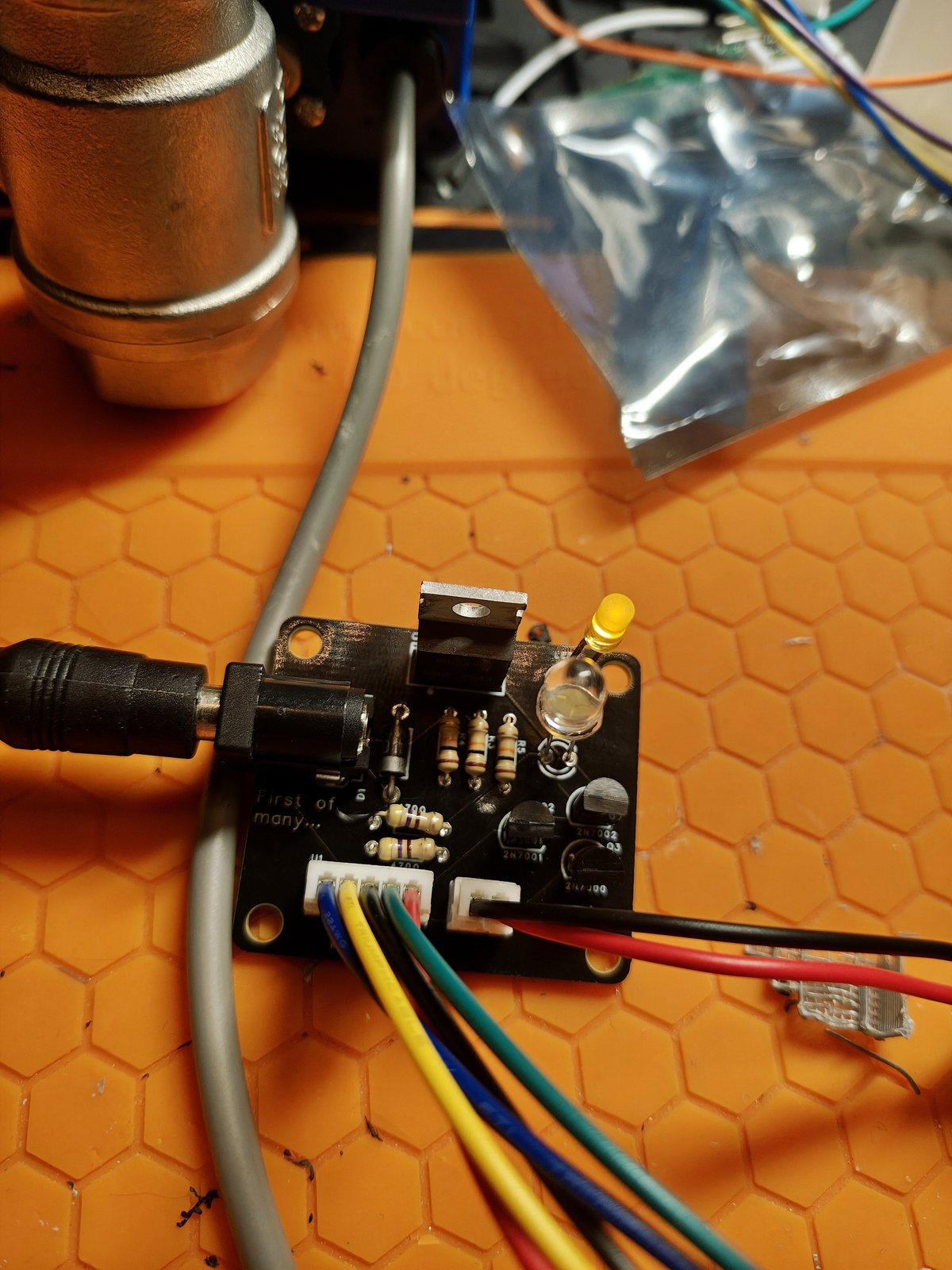 Close-up of the finished black PCB on an orange honeycomb mat. Every footprint is filled: the TO-220 IRF610 with its metal tab, three TO-92 2N7000 transistors, a row of three 10K gate resistors and two 470 ohm LED resistors, two indicator LEDs (a small yellow one and a larger clear dome), and the DC barrel jack on the left with a 12 V plug pushed in. Two white JST connectors carry the switch and the valve's wires — red, black, green, blue and yellow leads run off towards the bottom — and a grey valve cable curves up to the brass valve body at the upper left. 'First of many...' is on the silkscreen.